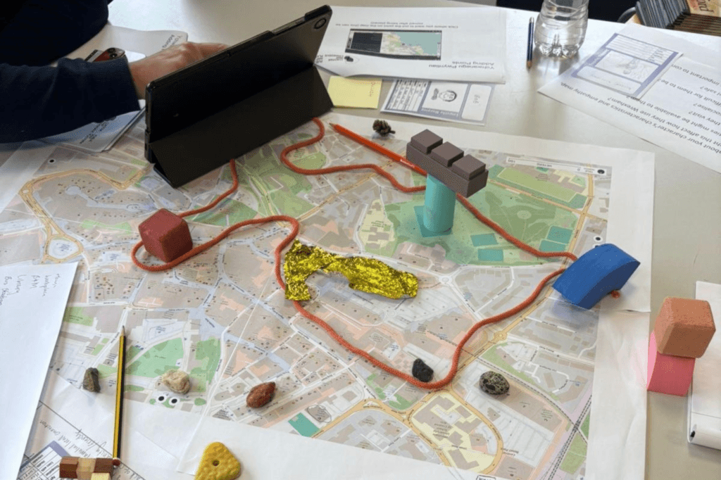 A map is laid out on a table with various objects and a tablet on top, suggesting a planning or discussion activity.