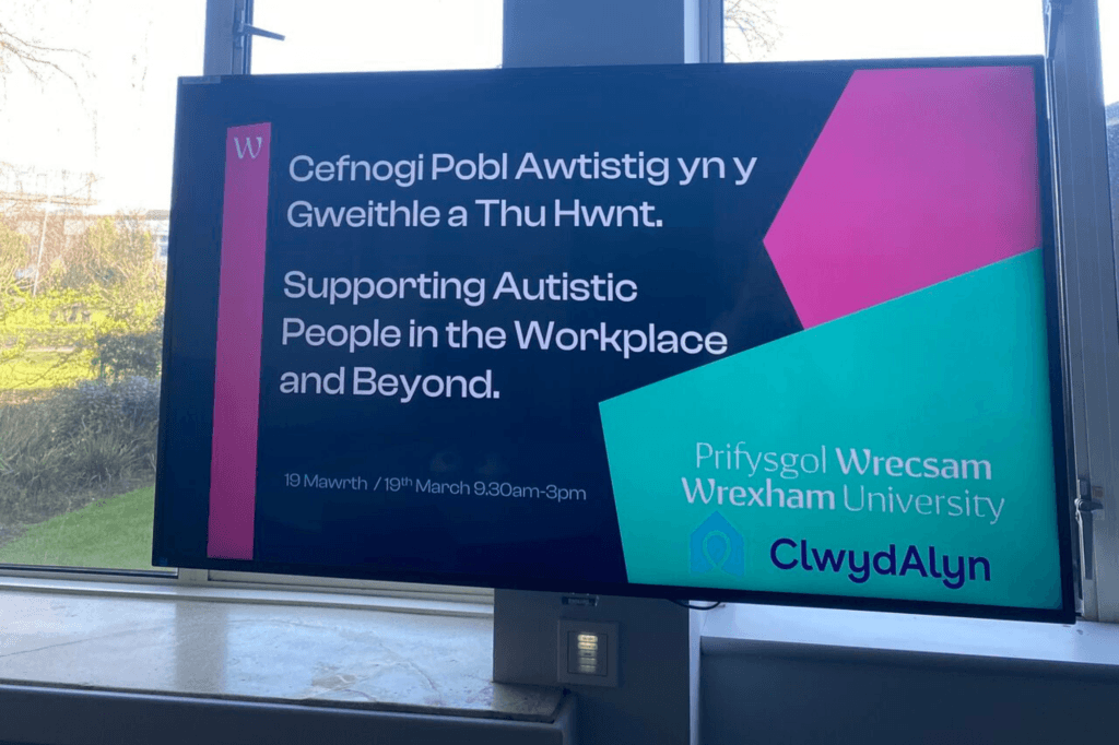 A digital screen displays information about supporting autistic people in the workplace. The text is in both Welsh and English, with the event date and time listed as March 10th, 9:30 AM to 3 PM. The screen also shows the logos for Wrexham University and ClwydAlyn.