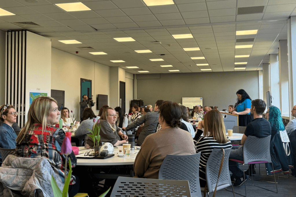 A speaker addresses an audience seated at round tables in a conference room. Some attendees are facing the speaker, while others are seen from behind.