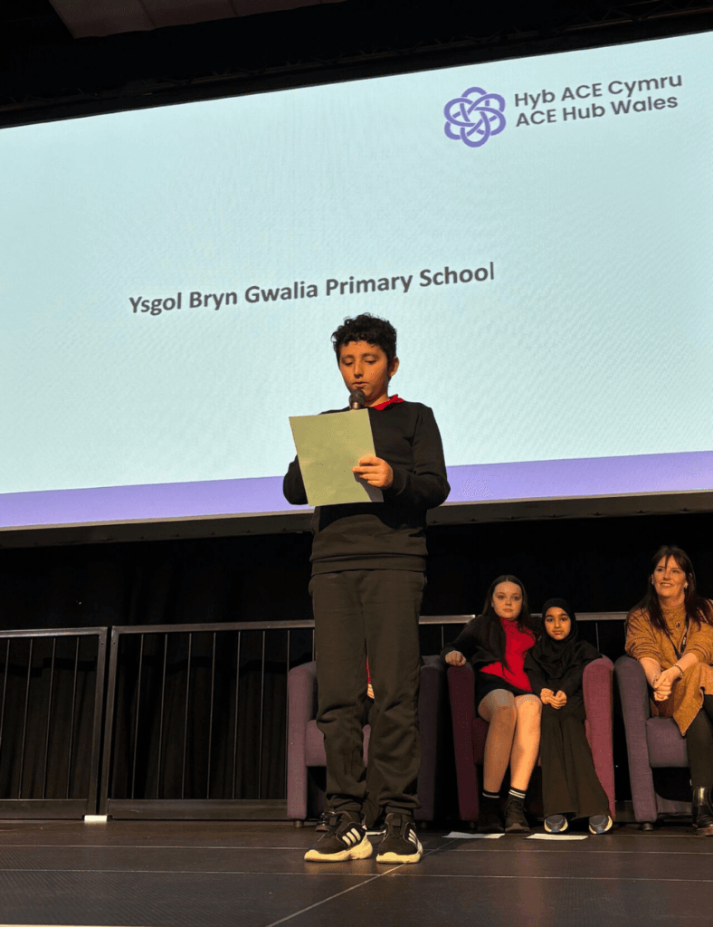 A student stands on a stage holding a paper and reading, with the text "Ysgol Bryn Gwalia Primary School" displayed on a large screen behind them. Other students are seated in chairs behind the presenter.
