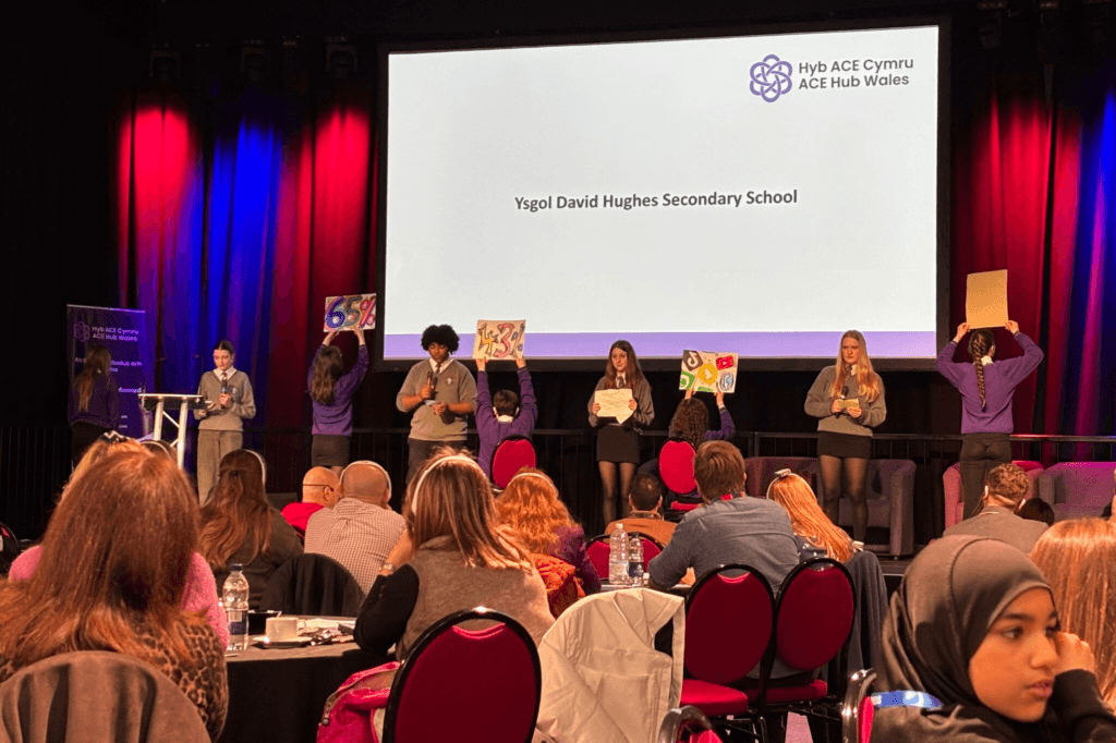 Students holding posters with statistics on on a stage in front of a large screen displaying "Ysgol David Hughes Secondary School"