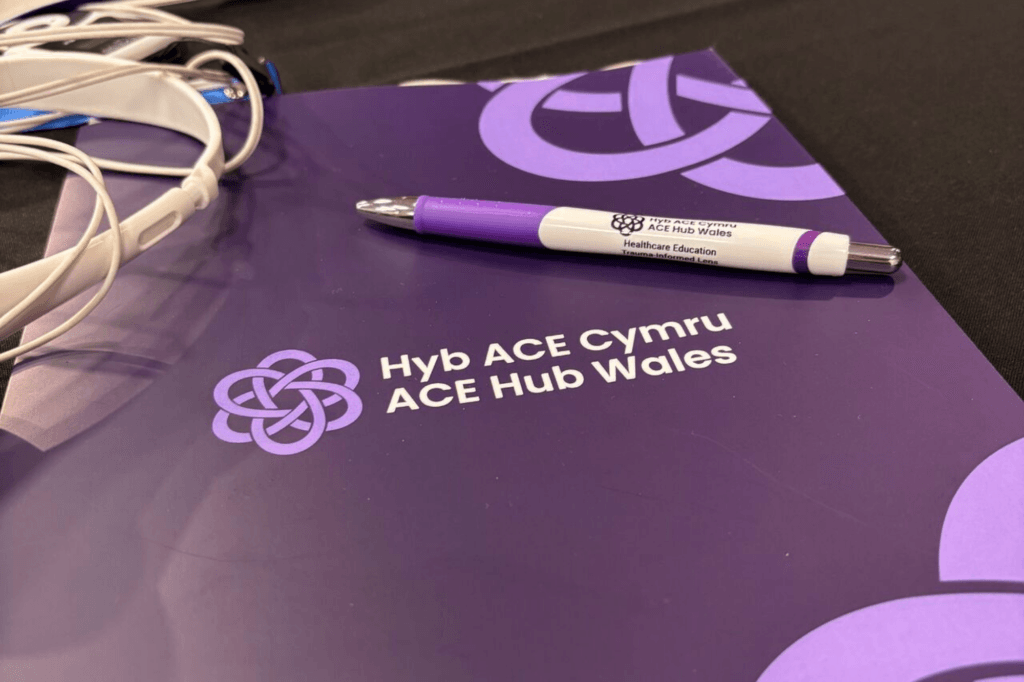 A purple folder with the text "Hyb ACE Cymru ACE Hub Wales" and a Celtic knot logo is on a table with lanyards and a pen.