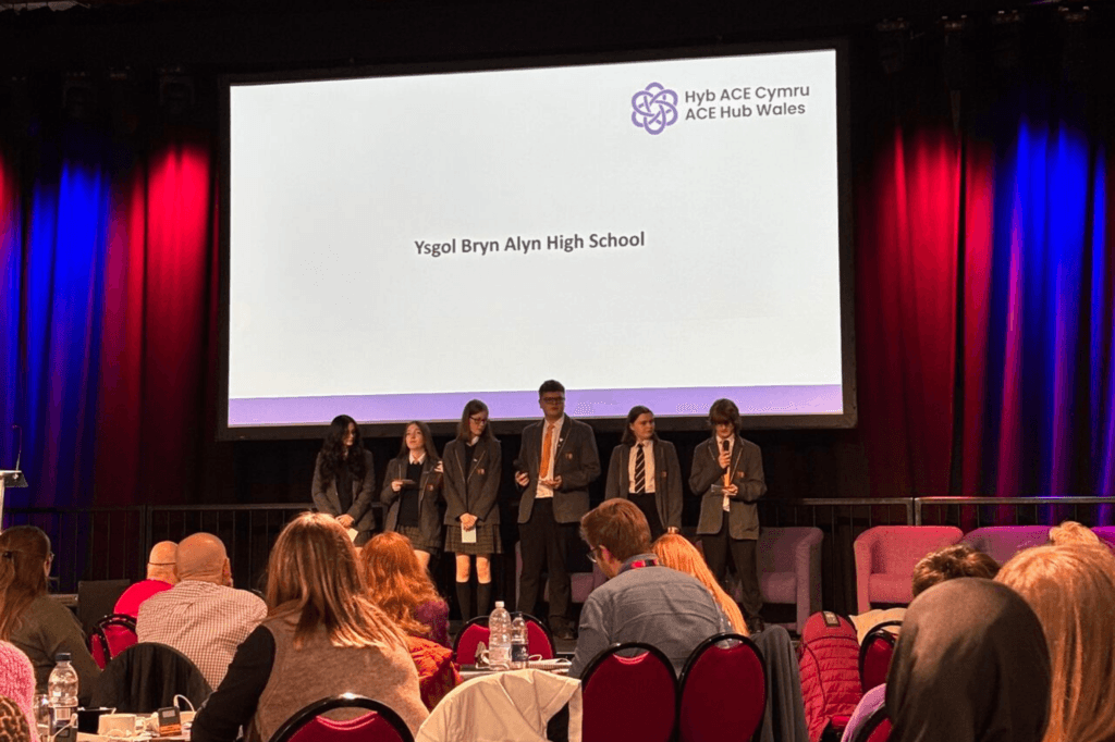 A group of students stand on a stage in front of a large screen displaying "Ysgol Bryn Alyn High School". The audience is seated in chairs in the foreground.